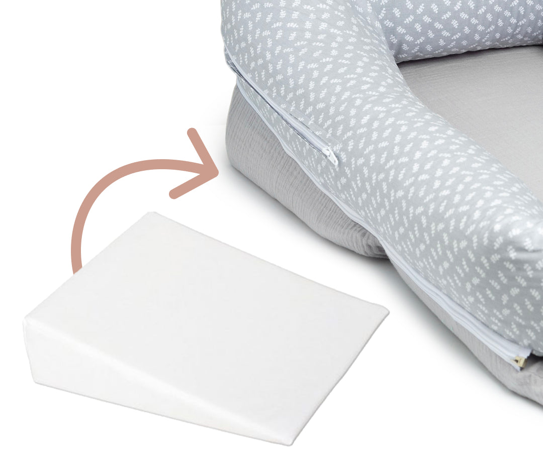 Un cojín para bebés gris con estampado blanco está abierto, mostrando una cuña de espuma blanca que se puede insertar en su interior. Una flecha marrón señala la cuña, indicando cómo debe colocarse en el cojín.